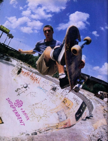 A skateboarder mid-air performing a trick at a graffiti-covered skate bowl under a bright blue sky with clouds. The skateboard is tilted vertically, showing a dark wood-grain top with light-colored wheels and black trucks.