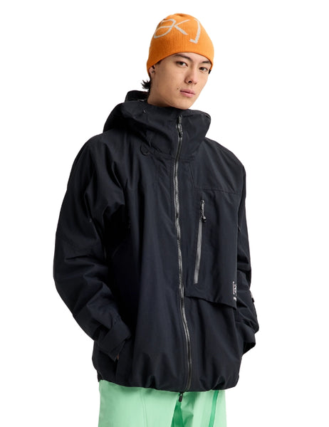 A model wearing a black hooded technical jacket with gray zippers, an orange beanie with a white logo, and mint green pants, set against a plain white background.