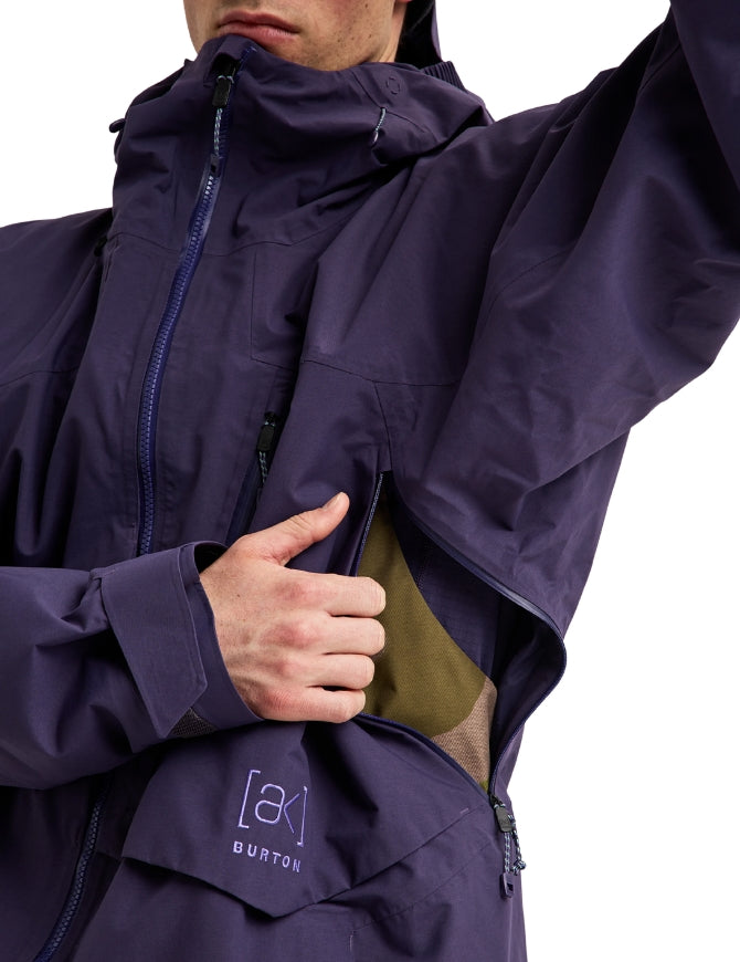 Close-up of a person wearing a dark purple Burton jacket, showing the chest pocket area with a hand opening the pocket to reveal an olive green lining. The Burton logo is visible on the lower front pocket.