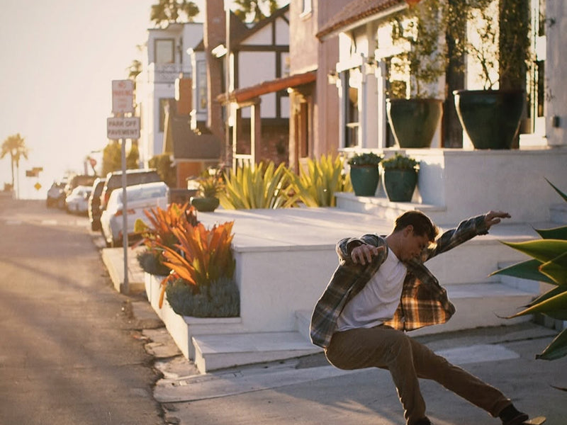 Person skateboarding on a sidewalk with residential buildings and palm trees in the background
