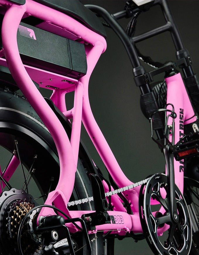 Close-up of a pink electric bicycle frame with black components, including the rear wheel, chainring, and handlebars, set against a dark studio background.