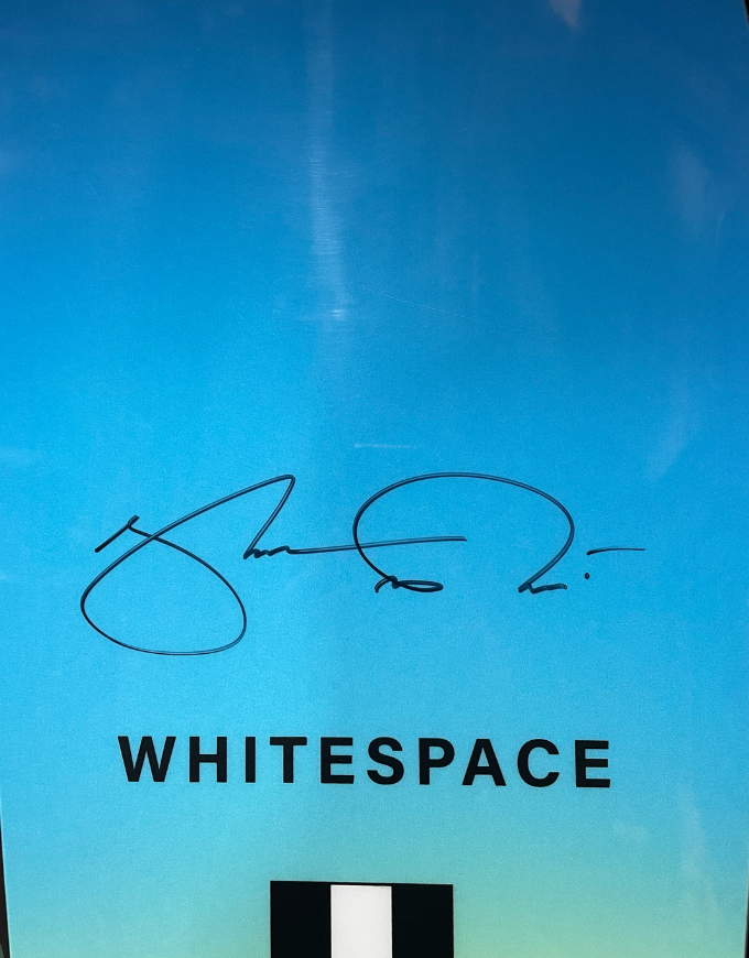 A blue gradient sign featuring a black cursive signature, the word 'WHITESPACE' in bold black uppercase letters, and a small black-and-white square at the bottom.