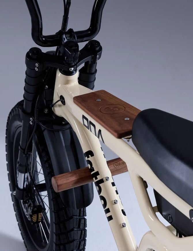 Close-up of a vintage-style bicycle frame in cream with black components, including a black seat, black handlebars, and black front tire. The frame features a cream-colored finish with black lettering and a brown wooden fuel gauge mount and handlebar mount.
