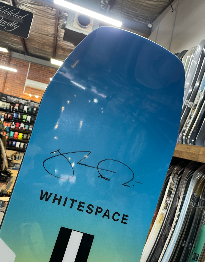 A close-up of a blue and green gradient snowboard featuring a black signature and the word 'WHITESPACE' printed in black, photographed inside a retail store.