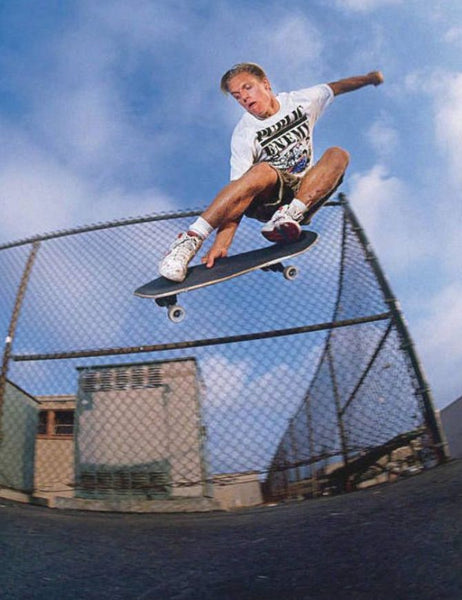 A skateboarder mid-air performing a trick in an outdoor skatepark, wearing a white graphic t-shirt, brown shorts, and white sneakers, with a chain-link fence and blue sky in the background.