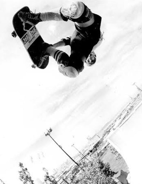Black and white action photograph of a skateboarder mid-air performing a trick, with a snowy outdoor background and a skateboard visible.