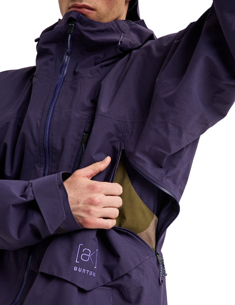 Close-up of a person wearing a dark purple Burton jacket, showing the chest pocket area with a hand opening the pocket to reveal an olive green lining. The Burton logo is visible on the lower front pocket.