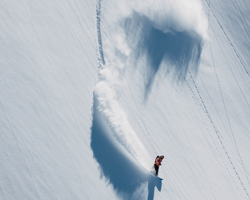 A snowboarder carving through deep powder snow, creating a dramatic spray of white snow and casting a long shadow on a steep, sunlit slope.