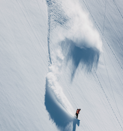 A snowboarder carving through deep powder on a snowy mountain slope, leaving a large spray of snow behind them. The scene is captured from above, with the skier appearing as a small figure near the bottom right.