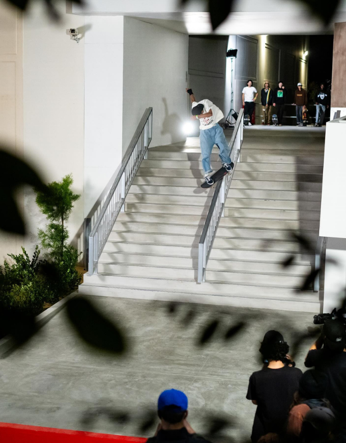 A skateboarder in a white t-shirt and light blue jeans slides down a wide concrete staircase at night, with a crowd of onlookers and photographers gathered at the bottom and top of the stairs.