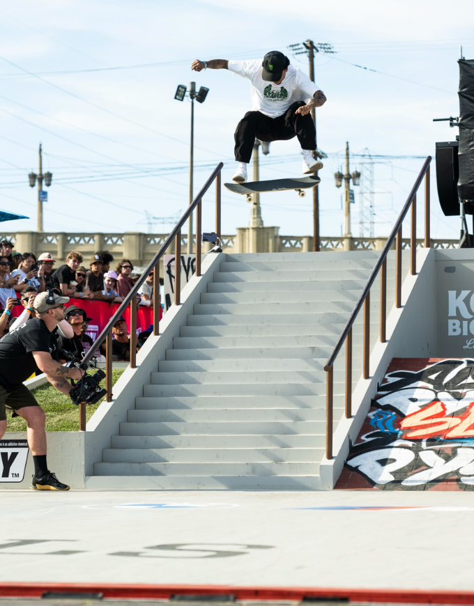 A skateboarder in a white t-shirt and black pants jumps a skateboard over a set of outdoor stairs, with a crowd of spectators and a photographer watching from the sidelines.