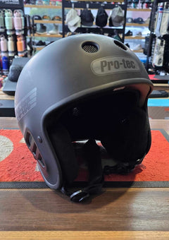 A matte dark gray Pro-tec ski helmet resting on a red mat on a wooden table inside a retail store.