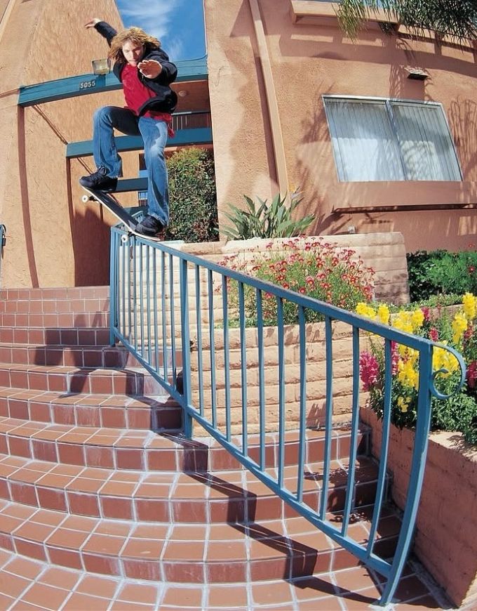 A person in a red and black jacket, blue jeans, and a black skateboard is performing a trick mid-air on a set of outdoor brick stairs with a blue metal railing. The background shows a stucco building with terracotta-colored walls and flower beds.