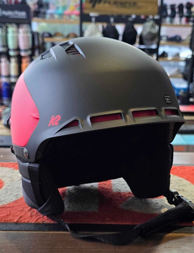 A matte black ski helmet with bright pink side panels and a pink K2 logo, resting on a patterned red and gray rug inside a retail store.