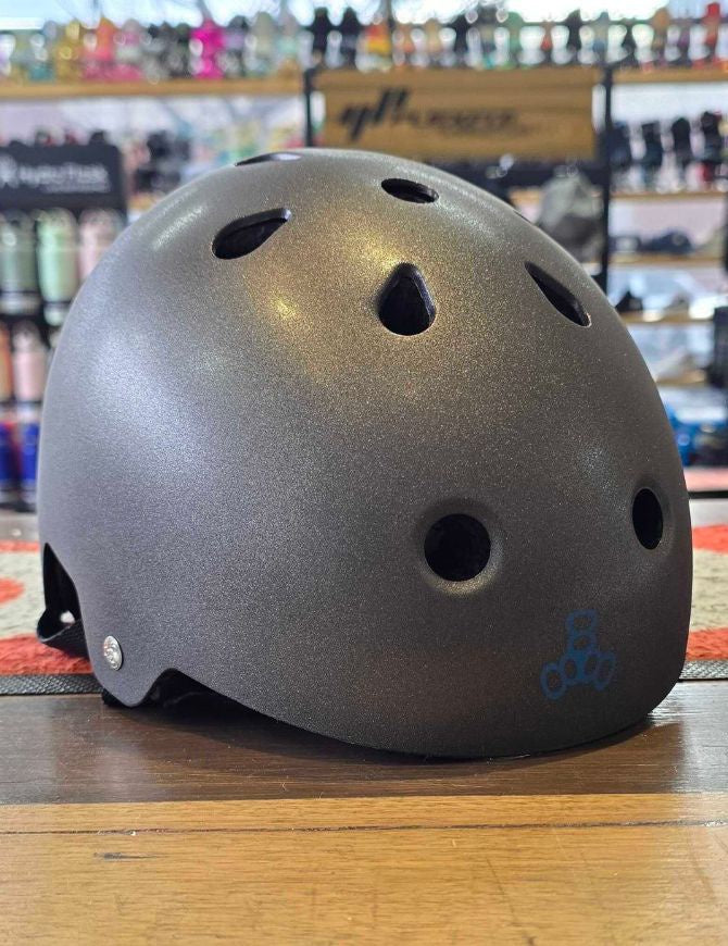 A dark metallic gray helmet with multiple ventilation holes and a small blue logo on the lower right, resting on a wooden table inside a store.